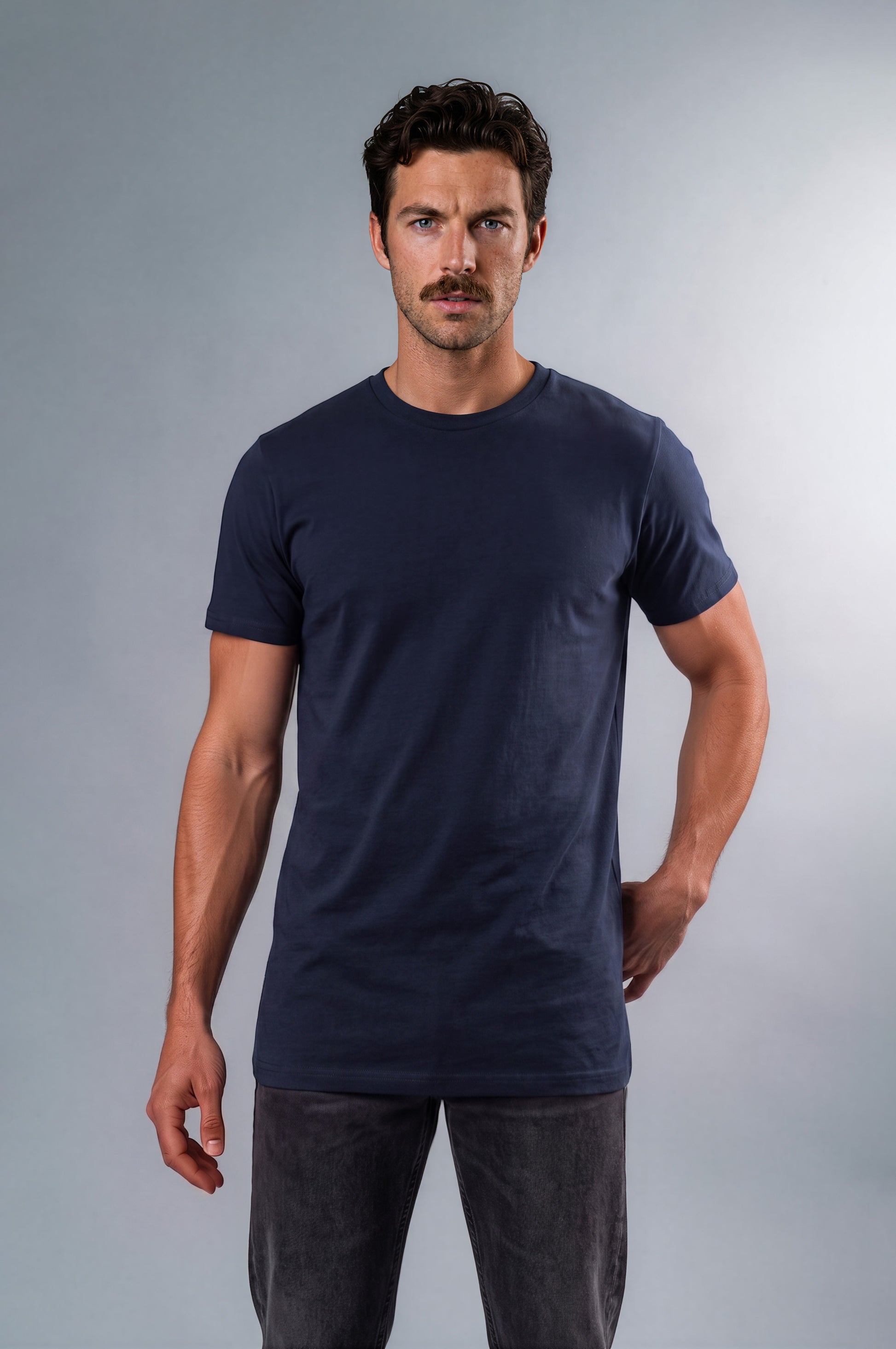 A man modeling a short-sleeve crewneck tall t-shirt in dark navy blue, paired with dark gray jeans, against a neutral gray studio background.